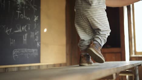 toddler boy walks away along wooden table in hut, dad holds him steady