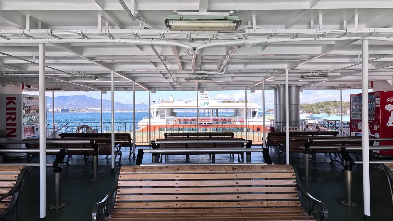 Interior of a JR ferry docked at Miyajimaguchi Port in Hiroshima, Japan, showing empty benches and a scenic view of Hiroshima Bay with a moored ferry in the background.