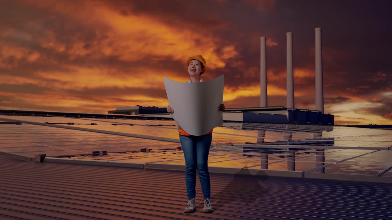 Ingegnere donna asiatica a figura intera con casco di sicurezza, che guarda un progetto nelle sue mani e si guarda intorno mentre è in piedi, ispezionando pannelli solari su un tetto al tramonto.