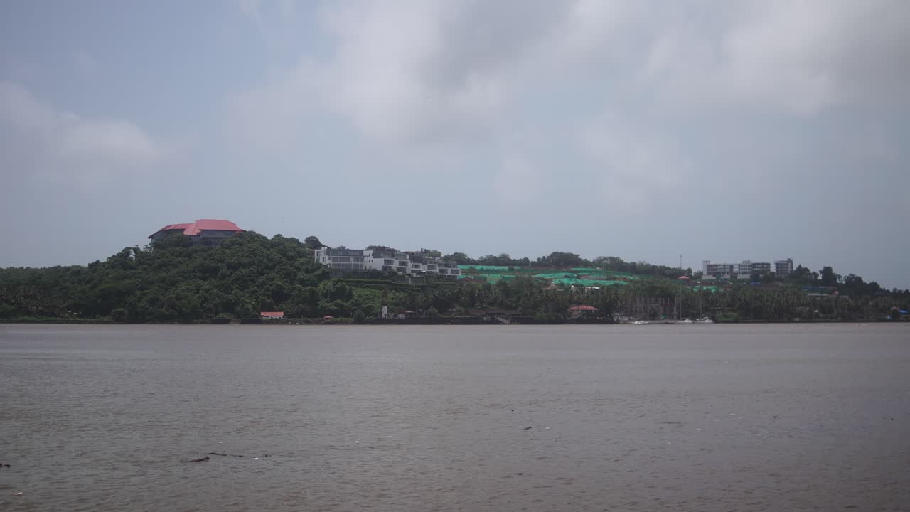 Mandovi River gliding through Goa, revealing waterfront landscape with verdant greenery, architectural structures under cloudy skies