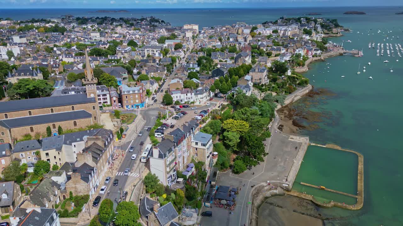 A drone pullback shot shows Notre-Dame d’Émeraude Church, the port, and the city of Dinard, with the sea and boats visible in the distance