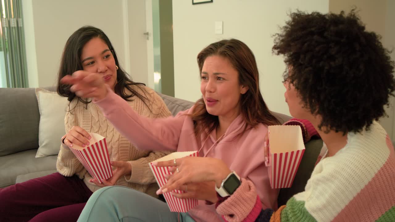 Diverse amiche reagiscono a un film fuori campo, afferrando confezioni di popcorn e indicando qualcosa su un divano grigio