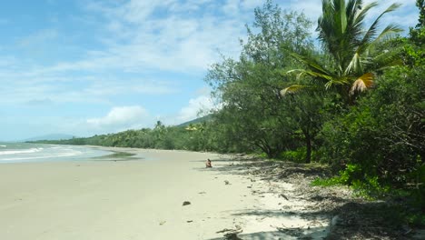 girl sits alone on a tropical beach with the ocean in front of her and the rainforest behind her
