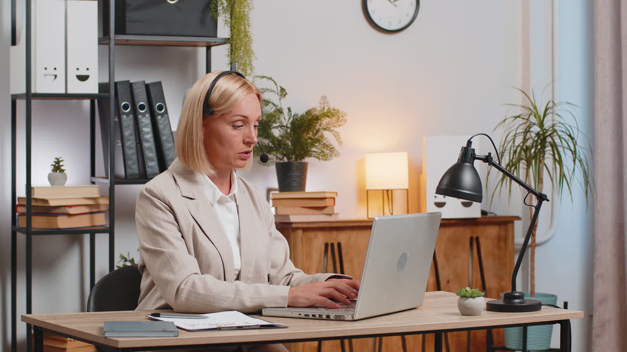 donna d'affari matura e sicura con le cuffie che parla su video con il portatile alla scrivania dell'ufficio a casa
