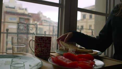 woman in pajamas prepares breakfast by the window, arranging bread and tomatoes on the table