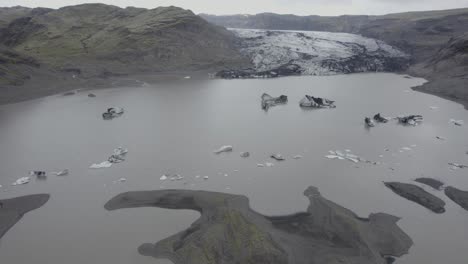 Aerial-view-overlooking-blackened-ice-blocks-on-the-Solheimajokull-glacier-lagoon---tracking,-drone-shot