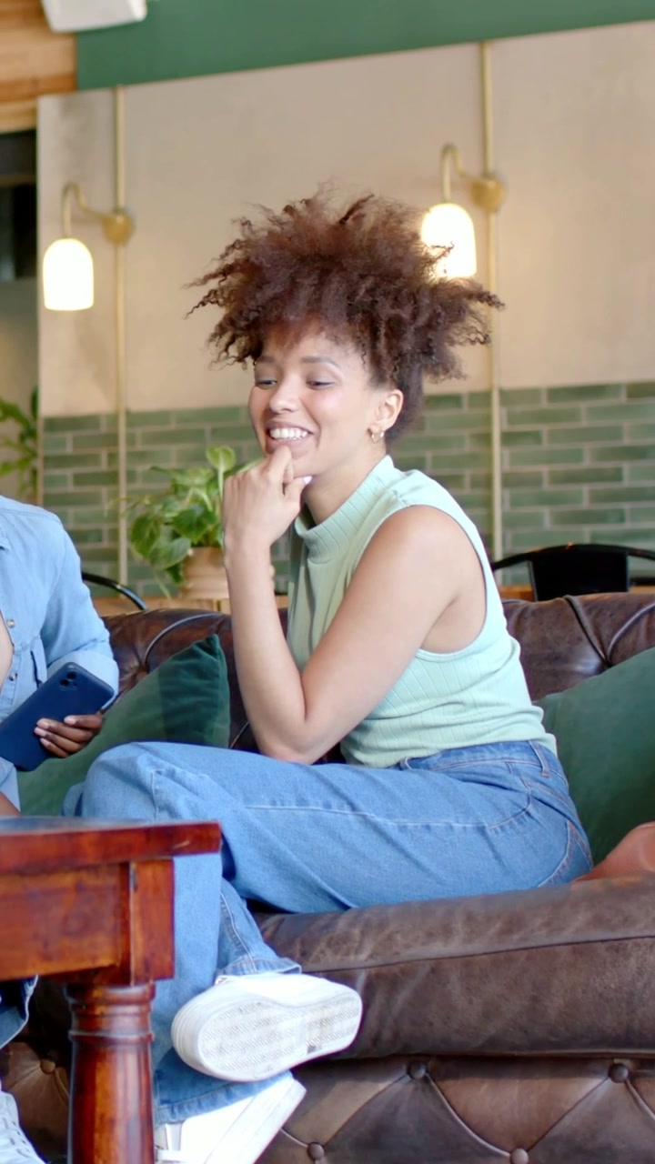 Vertical video: Smiling woman in mint top and jeans reaching for friend's phone on table in cafe