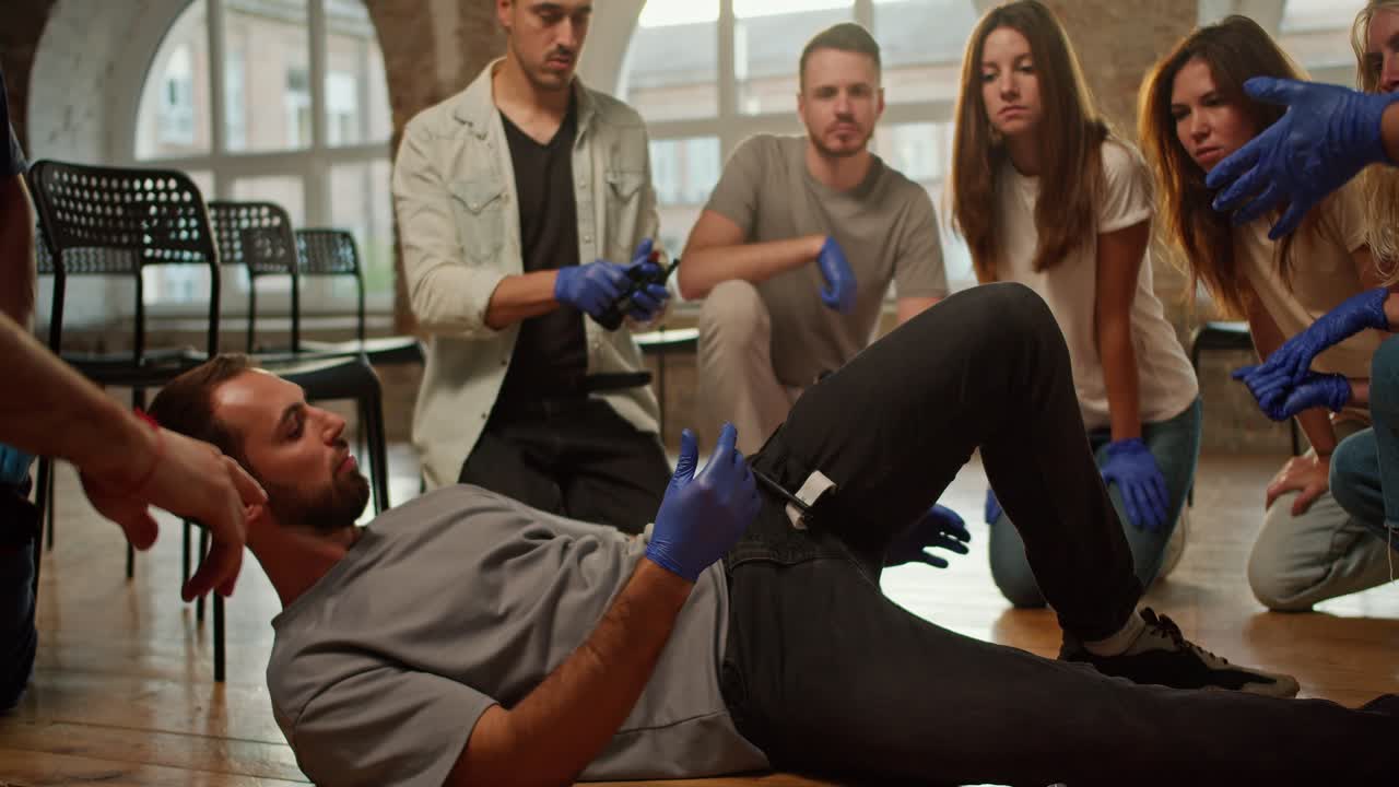 A bearded brunette man ties a medical tourniquet on her leg during a practical lesson in first aid and demonstrates it to others being trained