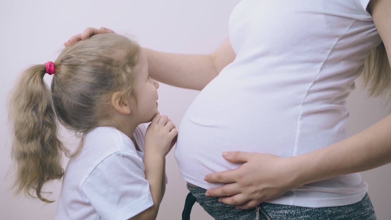 маленькая девочка нежно поглаживает беременную маму в комнате.