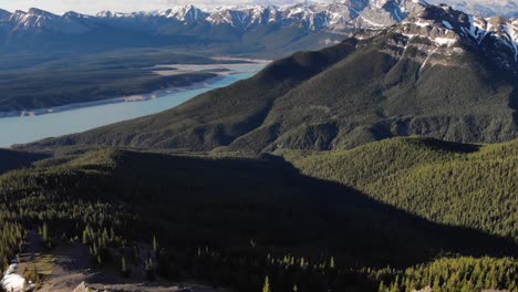 Aerial-tilt-up-shot-of-an-epic-view-of-a-valley-full-of-green-trees-with-a-mountain-range-with-rocky-snowy-peaks-in-the-background,-conservation-concept