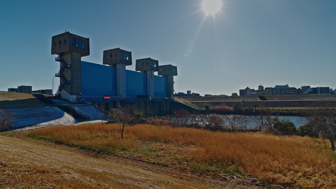 Uno scatto paesaggistico grandangolare della moderna Chiusa Blu di Iwabuchi a Tokyo. La scena presenta i cancelli industriali blu e le torri in cemento posti contro un vasto argine erboso sotto un cielo luminoso e sereno.