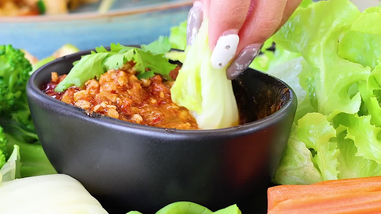 A hand dips fresh vegetables into a spicy chili sauce, surrounded by vibrant greens and herbs.