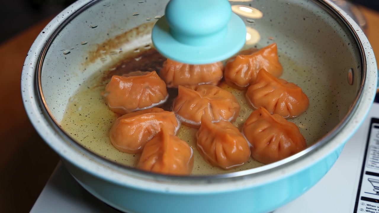 Bidikan jarak dekat tangan meletakkan tutup kaca ke wajan biru muda untuk menutupi pangsit oranye yang sedang digoreng. Dim sum Asia yang lezat itu dimasak dalam minyak di atas kompor portabel di dapur rumah modern dalam ruangan