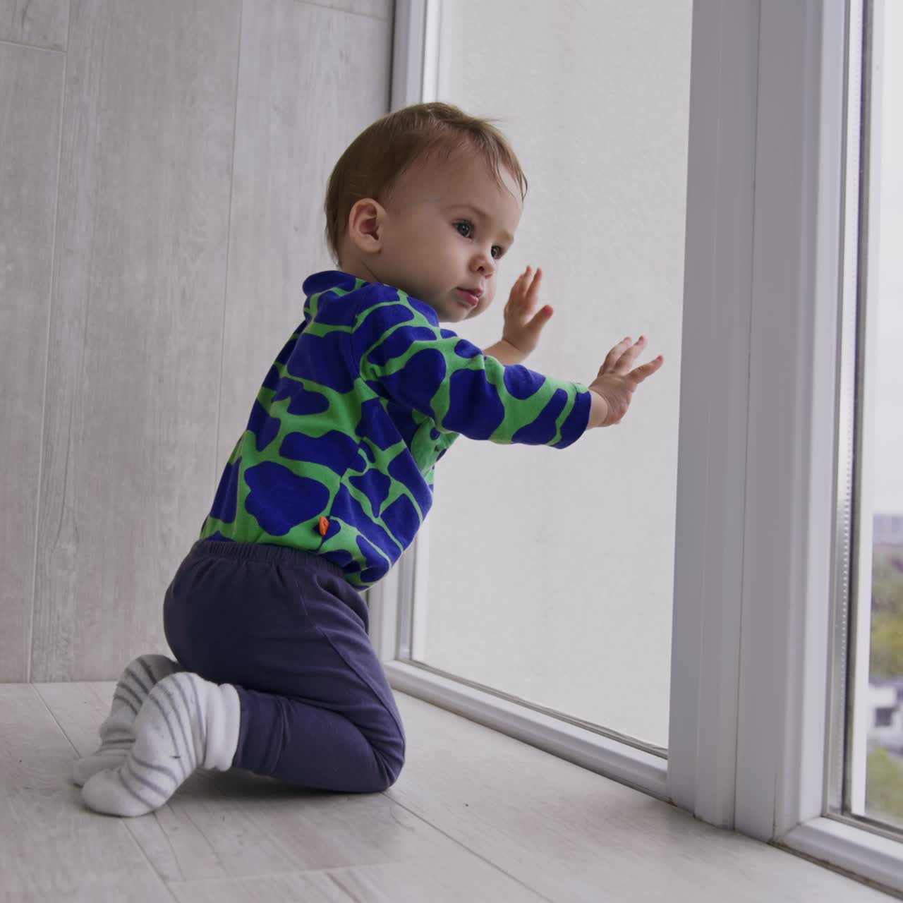 Caucasian baby boy playing on the floor near the big window. Toddler banging by the window with his hands and than distracts on something else
