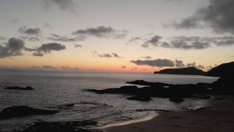 Spectacular-sunset-flight-above-Calheta-sandy-beach-toward-beautiful-ocean-sea-water,-rock-outcroppings,-silhouette-mountain-range-and-golden-hour-colorful-sky,-Portugal,-overhead-aerial-approach