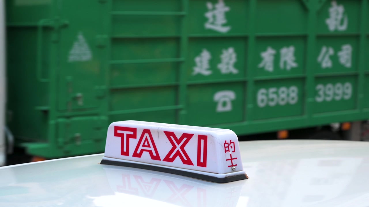 Un'insegna del taxi è sul cofano del veicolo mentre pedoni e traffico sono visibili sullo sfondo.