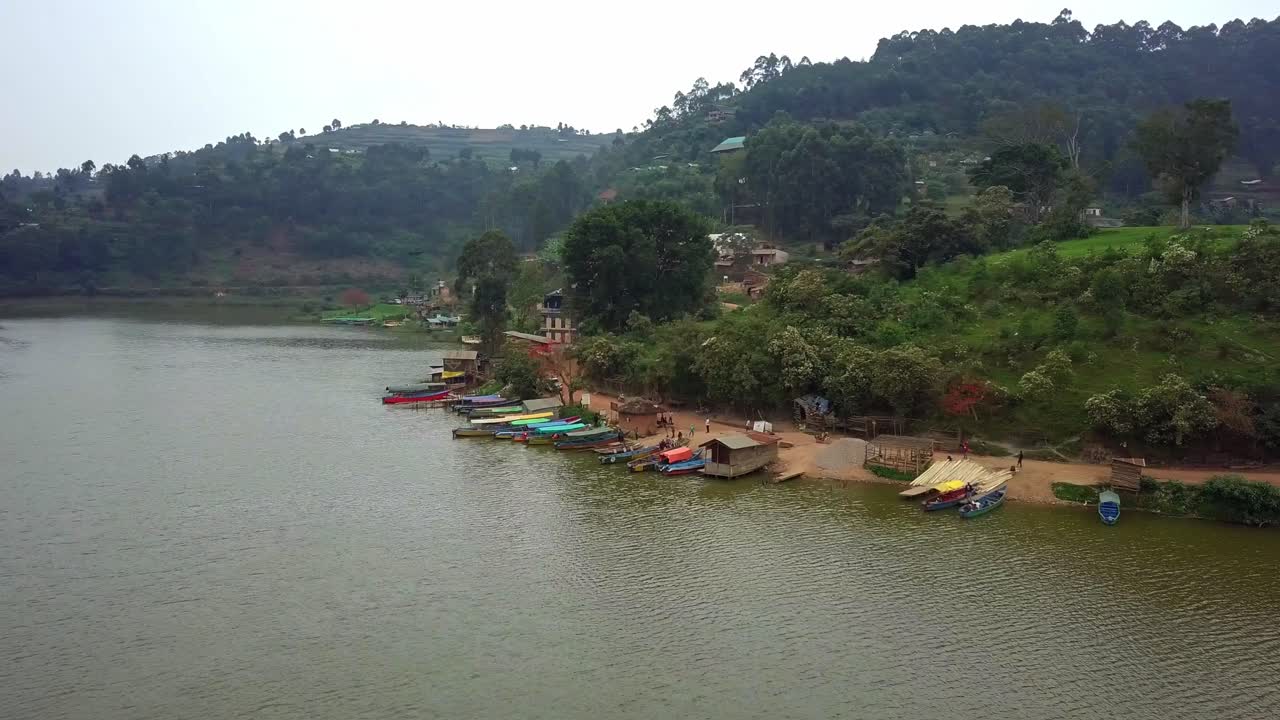 Drone orbiting view of docked boats in Rutinda on Lake Bunyonyi, Uganda, showing shoreline activity and terraced hills in a tranquil lakeside setting