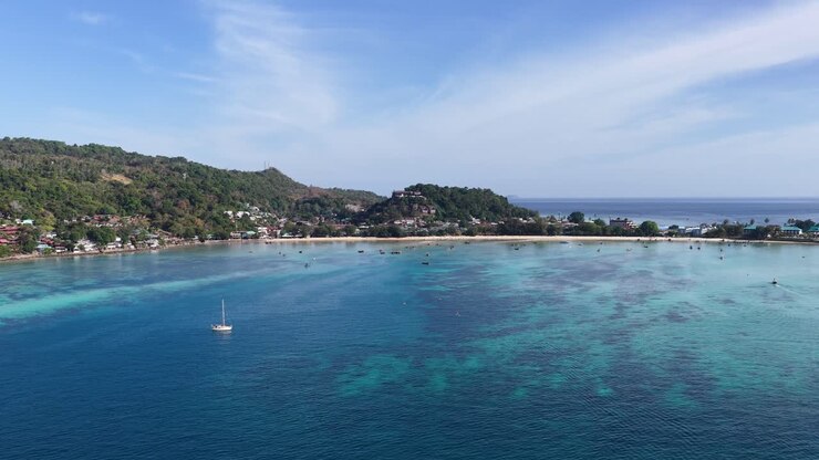 Ampia ripresa aerea con drone di una baia tropicale con barche a vela, acqua turchese e lussureggianti colline verdi.