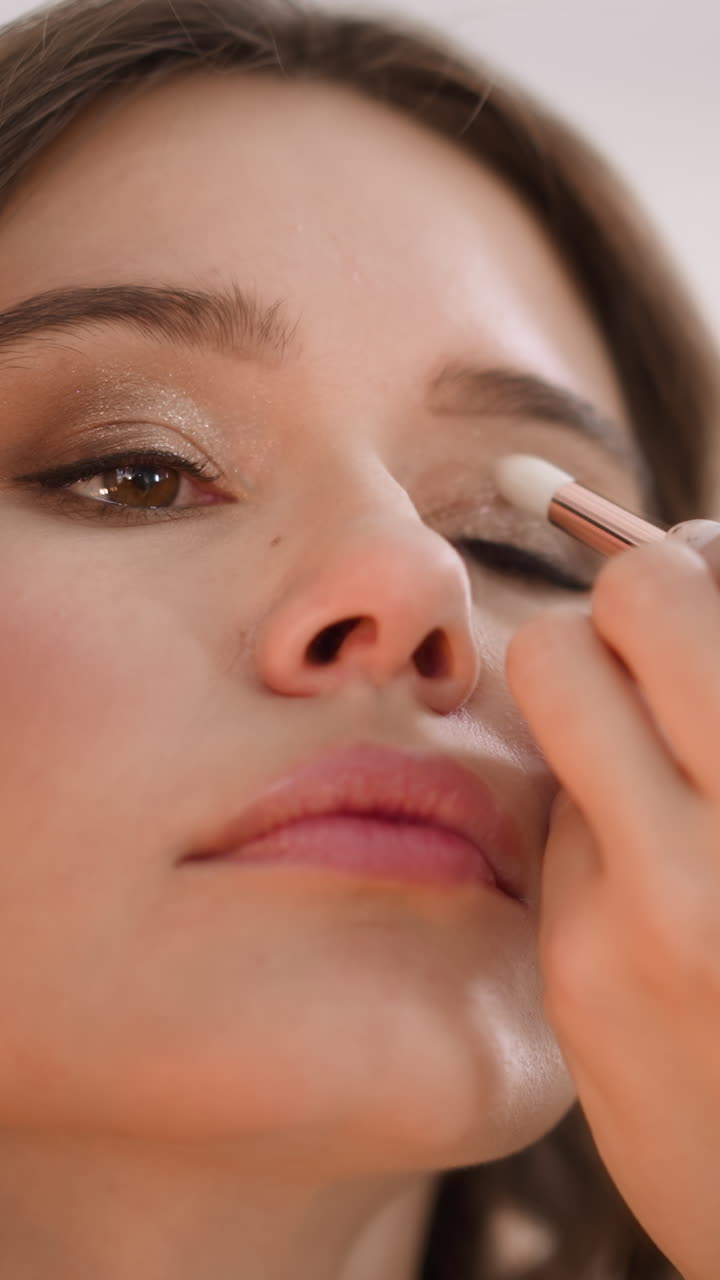 Focused woman applies glitter shadow on eyelid with applicator looking in mirror. Lady prepares for birthday party of friend doing makeup closeup