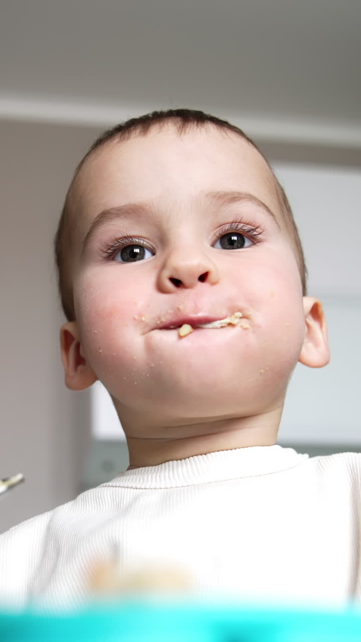Beautiful Caucasian toddler eating funnily from spoon. Some food stuck around kid's mouth. Low angle view. Vertical video
