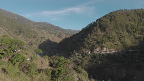 Un-Pueblo-En-Un-Valle-Cubierto-De-árboles-Verdes,-Montañas-Y-Cielo-Azul,-Nubes-Claras-En-El-Fondo,-Antena-Acercándose-Y-Sobrevolando