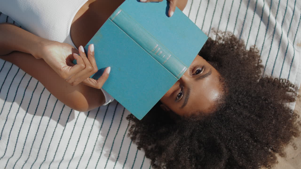 Primo piano di ragazza in spiaggia che legge un libro sdraiata su una coperta. Donna sorridente si rilassa (video verticale).