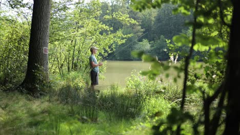 Un-Pescador-Masculino-Se-Encuentra-En-La-Orilla-De-Un-Lago-Nublado-Entre-Hierba-Verde-Y-árboles-Pescando-Con-Caña-De-Pescar,-Pan