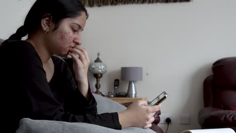 A-female-teenager-sits-in-the-living-room,-scrolling-through-her-phone's-touchscreen-while-seated-on-a-sofa-chair,-showcasing-the-integration-of-home-multimedia-usage-in-daily-life