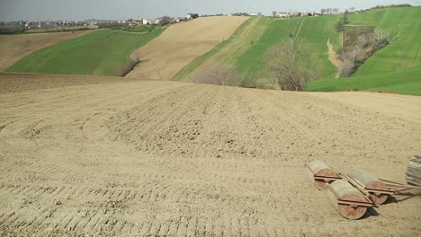 Primer-Plano-De-Un-Pequeño-Tractor-De-Orugas-Rojas-Tirando-De-Un-Sistema-De-Laminación-Triple-Preparando-La-Tierra-Antes-Del-Proceso-De-Siembra-En-El-Campo-Italiano-Con-Un-Pequeño-Pueblo-Escénico-En-El-Fondo