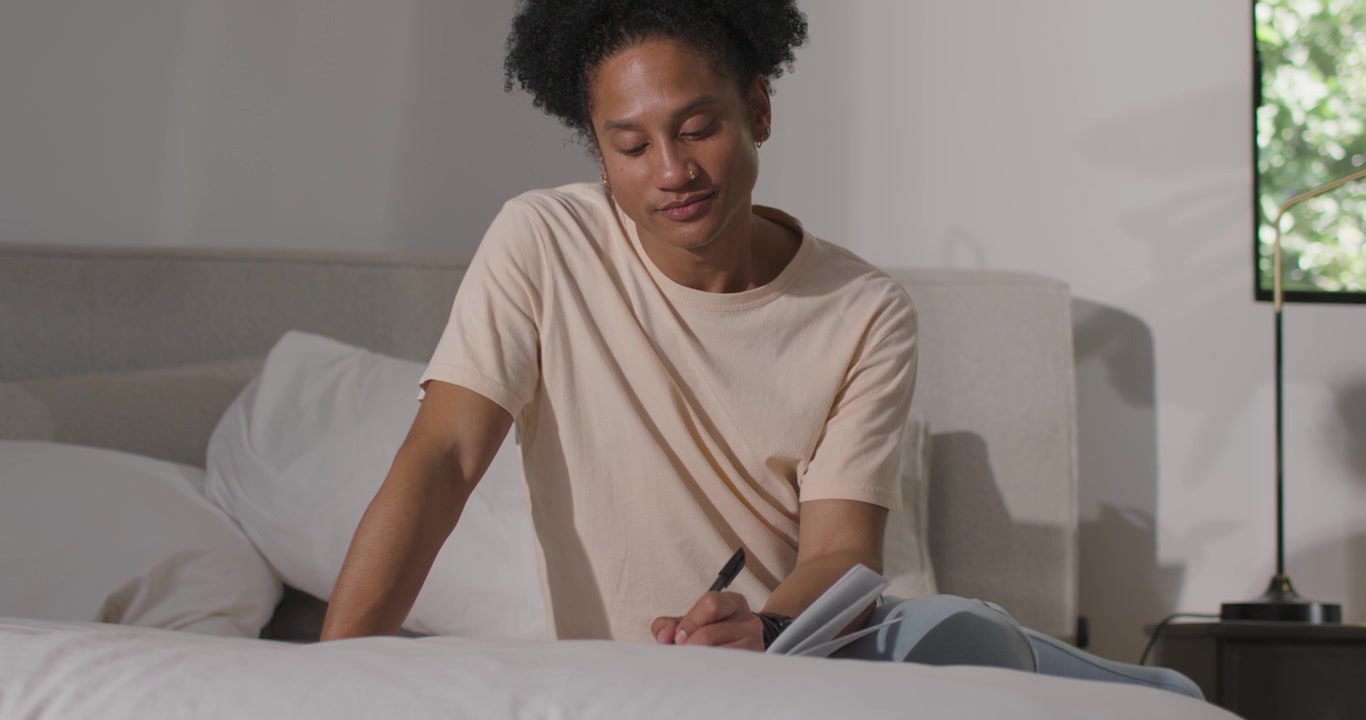 Uomo adulto che guarda un quaderno a spirale, allunga la mano con una penna e scrive per prendere appunti sul letto.