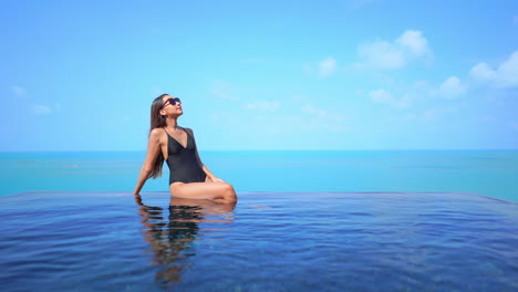 A-contrast-in-blues,-a-woman-in-a-bathing-suit-sits-on-the-edge-of-an-infinity-edge-pool-ocean-in-the-background