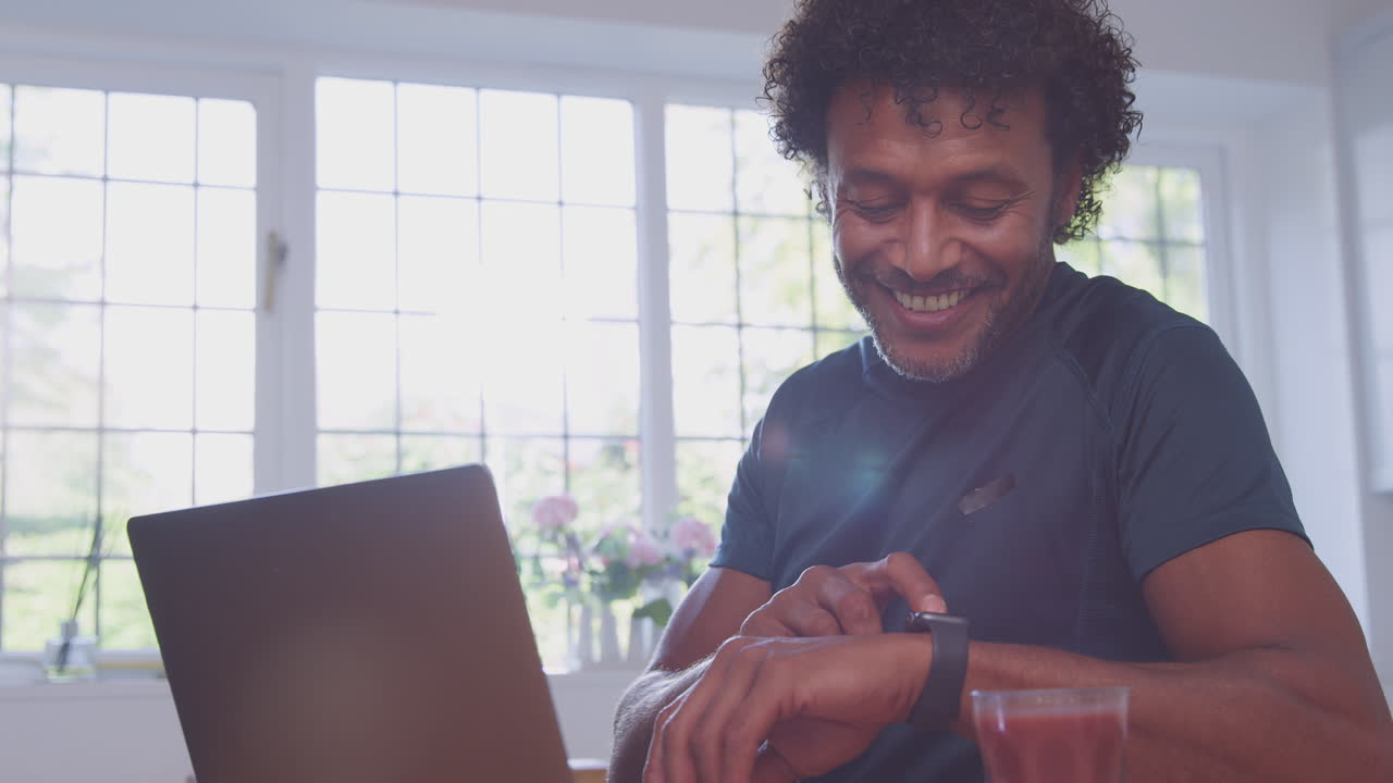 Mature Man Wearing Fitness Clothing At Home In Kitchen Logging Activity On Laptop Computer