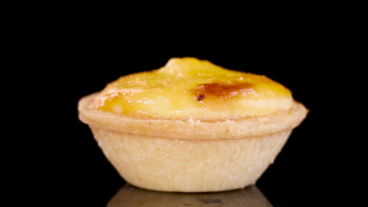 A single egg tart rotates smoothly on a glossy black surface under bright studio lighting, highlighting its golden crust and creamy filling in a controlled environment