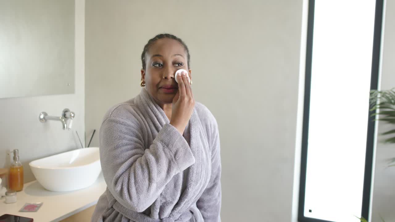African American woman reaching for cotton pad, cleansing skin at bathroom vanity, copy space