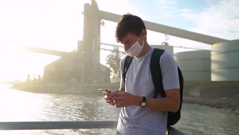 Hombre-Con-Mascarilla-Y-Mochila-Usando-Un-Teléfono-Inteligente-Cerca-Del-Puerto-En-Ingeniero-White,-Buenos-Aires,-Argentina---Plano-Medio,-Cámara-Lenta