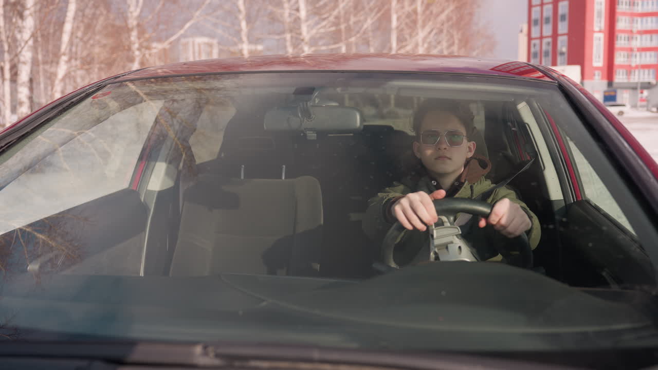 teenage boy in winter jacket lifts sun visor inside car, seen through front windshield with bare trees and snowy outdoor background, steering with one hand while adjusting visor with the other