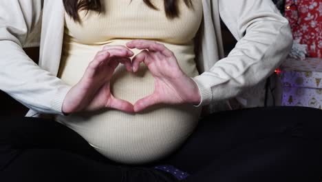 pregnant woman make heart shape with palm fingers near christmas presents