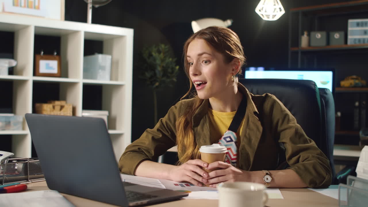 Ragazza sorridente che parla online in un ufficio buio. Donna impegnata che beve caffè al lavoro.