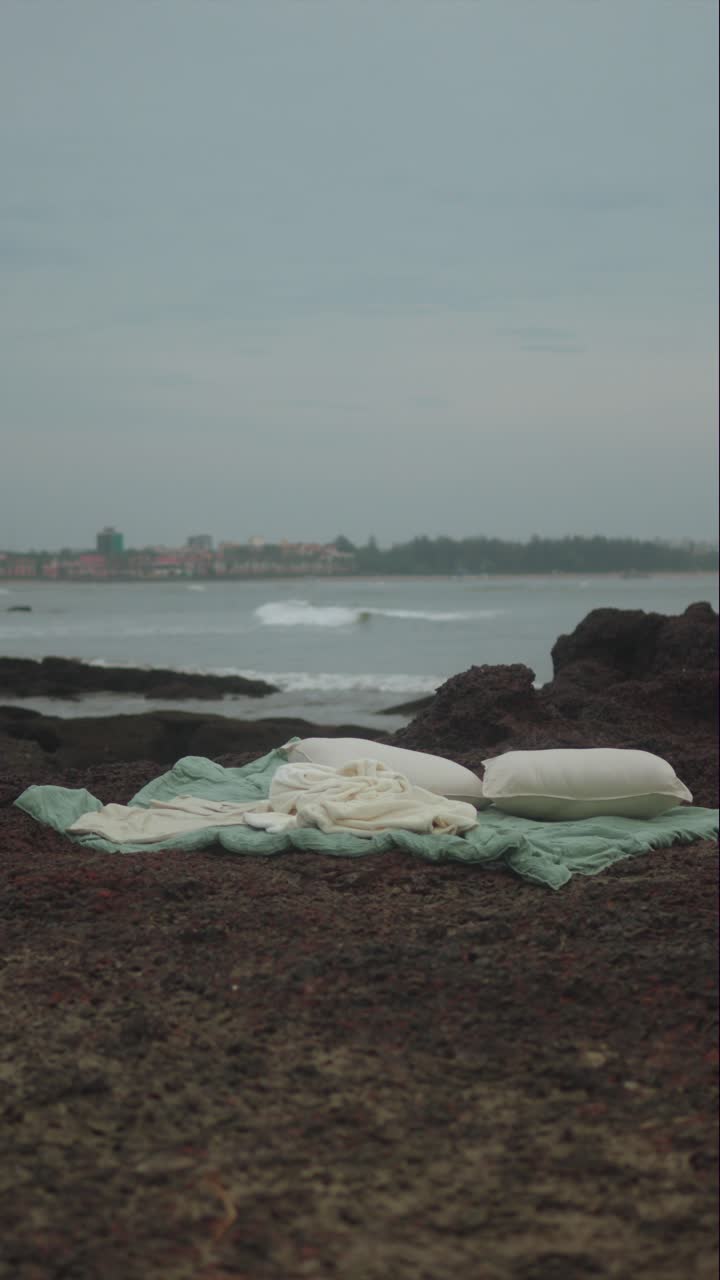 Vertical artistic composition of soft bedding and pillows placed on a rocky beach by the sea, blending comfort and wilderness in a poetic coastal scene
