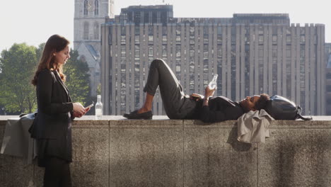 Two-millennial-female-friends-relaxing-at-the-Thames-embankment-holding-smartphones-and-talking,-one-lying-on-the-wall,-backlit,-close-up