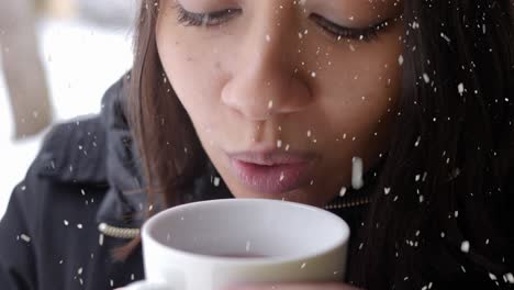 Asian-woman-blowing-wind-to-cup-of-tea-during-cold-winter-day-while-snowing,-close-up-view