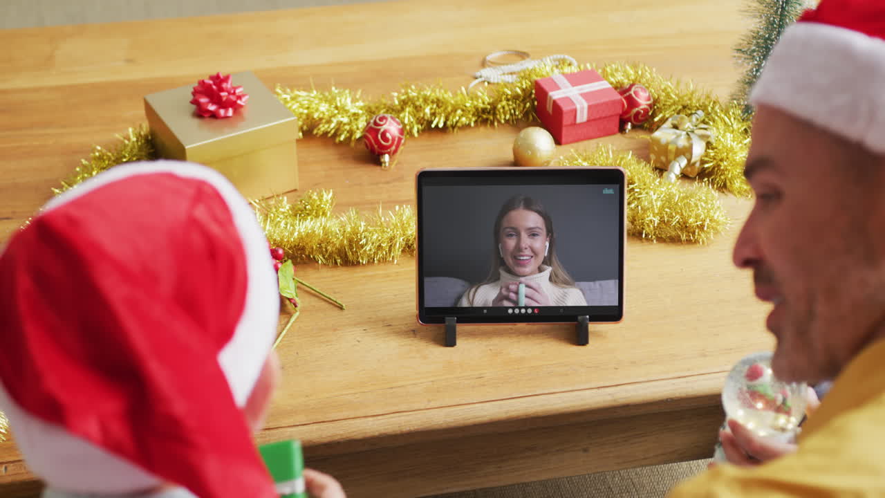 padre y hijo caucásicos con sombreros de santa usando una tableta para una videollamada de navidad con una mujer en la pantalla