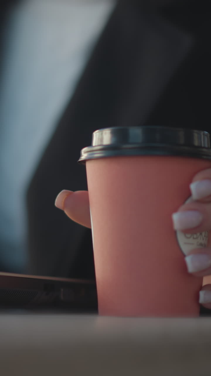 primer plano de una computadora portátil en la mesa con la mano en un abrigo negro, uñas pulidas, dejando caer una taza de café en la superficie de la mesa, alguien sentado, fondo borroso con un restaurante al aire libre