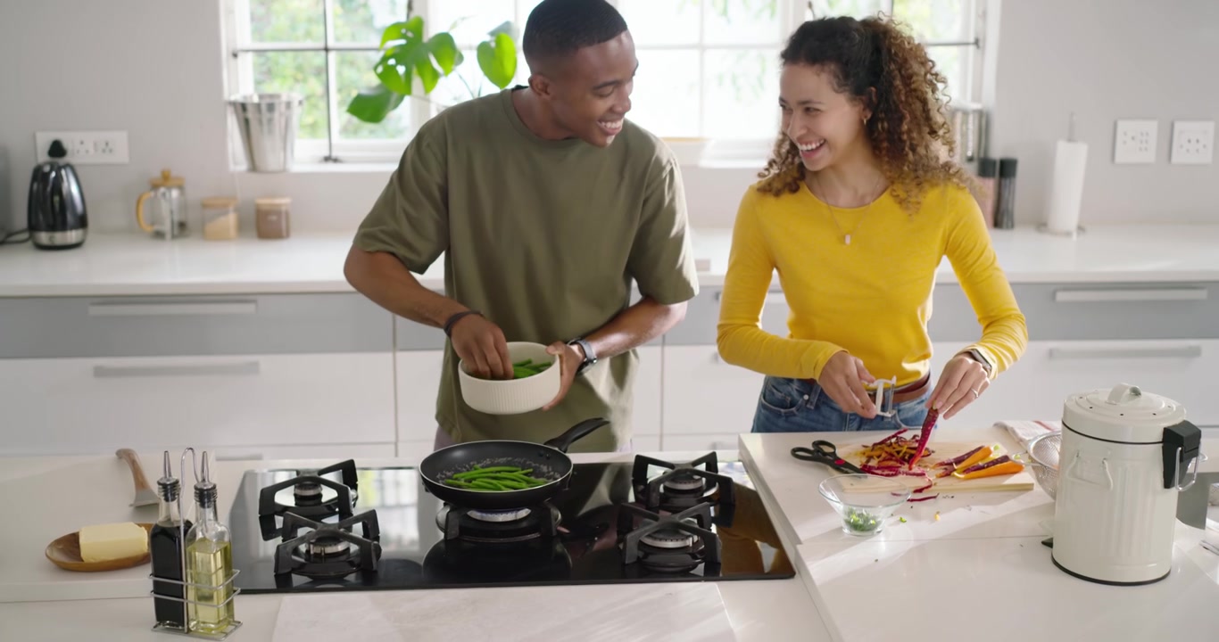 Casal diverso começando a refeição na ilha, homem pegando verduras da tigela para a panela enquanto a mulher pica.