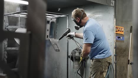 Blue-collar-worker-spray-painting-metal-with-powder-coating-in-an-industrial-room,-slow-motion