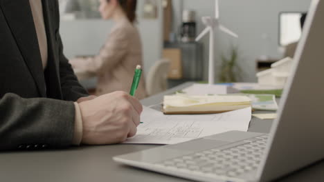 close up view of unrecognizable man writting on blueprint sitting at desk, then he stands ap and looks the papers