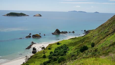 Aerial-panning-shot-of-tropical-sandy-beach,-blue-ocean,-green-meadow-on-mountain-and-islands-in-background---Sunny-day-at-Te-Whara-Track-in-New-Zealand