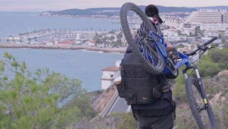 Traveller-carries-mountain-bike-on-one-sholder-down-hill-as-bendy-road-down-below-leads-to-the-coastal-city