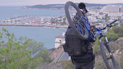 traveller carries mountain bike on one sholder down hill as bendy road down below leads to the coastal city