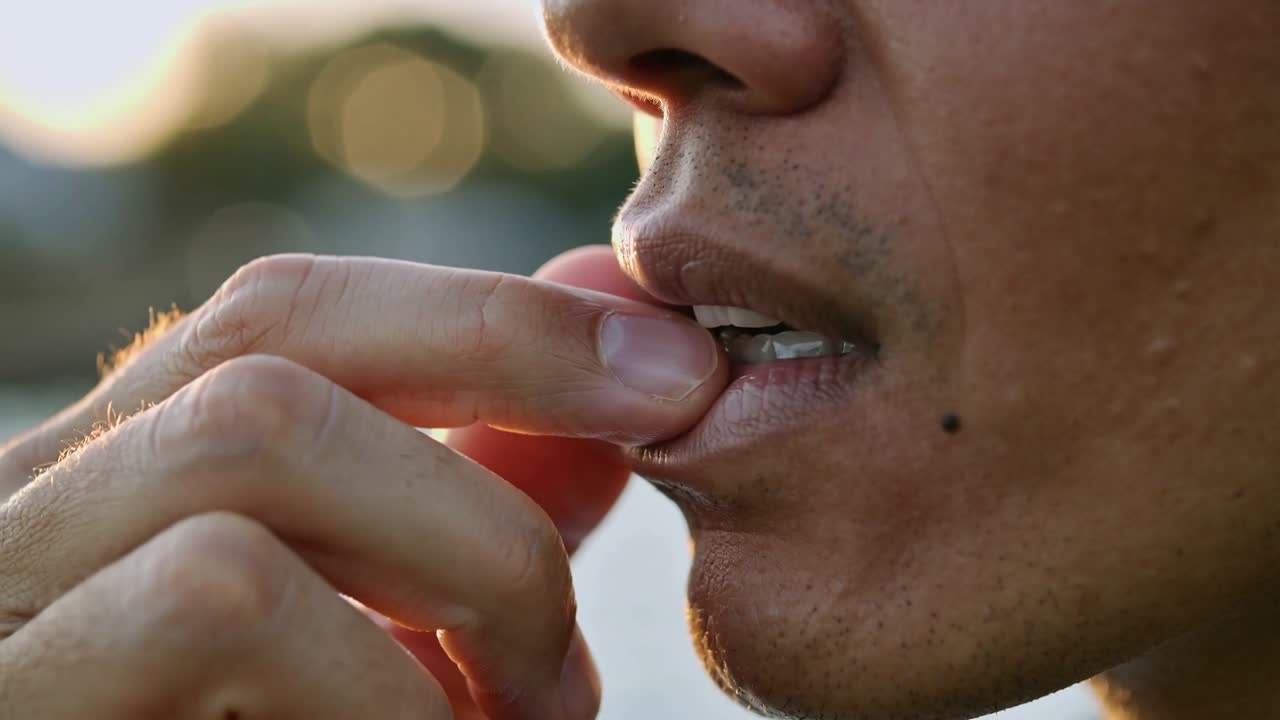 Pressing stubbled man's right fingertip against lower lip, checking chapped lip under late sunlight